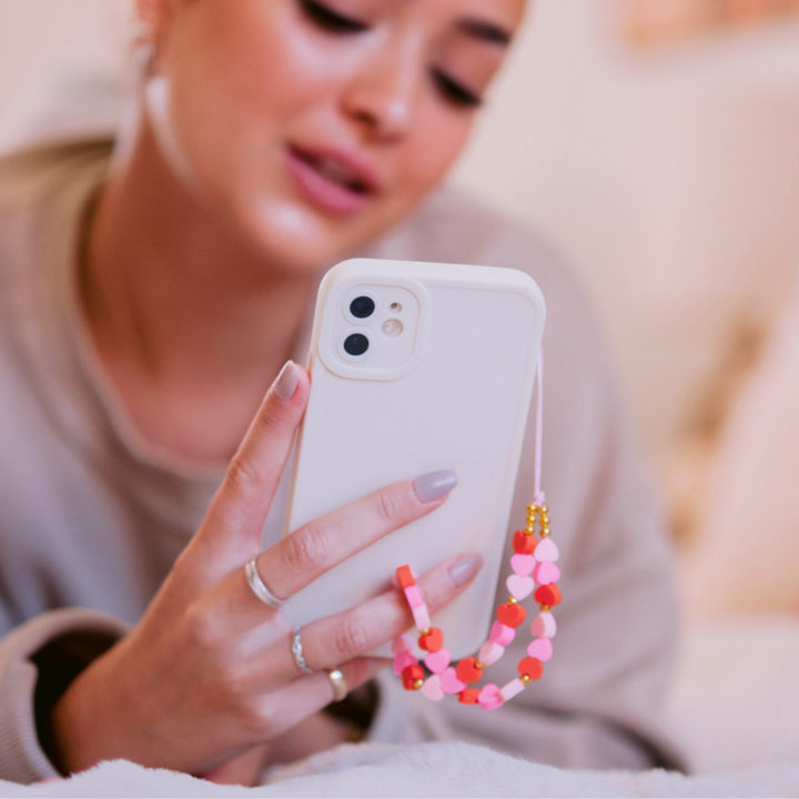 A person with light nail polish holds a white smartphone adorned with the Coconut Lane Beaded Phone Strap - Hearts in pink and orange. The person is wearing a beige top and has their face slightly out of focus in the background.