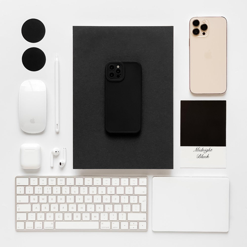 A neatly arranged workspace with a black iPhone in a Coconut Lane NAKD Phone Case - Midnight Black on a dark notebook, a gold iPhone, a white keyboard, Magic Mouse, AirPods, white touchpad, stylus, two black circles, an AirPods case, and a "Midnight Black" color sample card—all on a white surface.