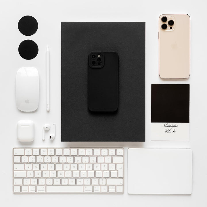 A neatly arranged workspace with a black iPhone in a Coconut Lane NAKD Phone Case - Midnight Black on a dark notebook, a gold iPhone, a white keyboard, Magic Mouse, AirPods, white touchpad, stylus, two black circles, an AirPods case, and a "Midnight Black" color sample card—all on a white surface.