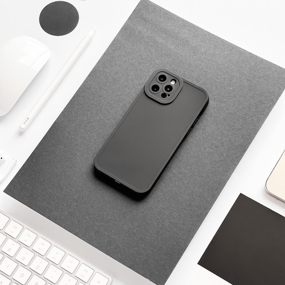 A black smartphone featuring the NAKD Phone Case in Midnight Black by Coconut Lane sits on a gray sheet of paper on a white desk. Surrounding the phone are various items, including a white keyboard, a pen, a mouse, and some papers. The image showcases a minimalistic and monochromatic color scheme.