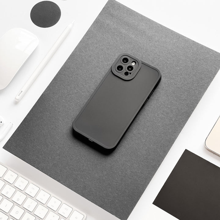 A black smartphone featuring the NAKD Phone Case in Midnight Black by Coconut Lane sits on a gray sheet of paper on a white desk. Surrounding the phone are various items, including a white keyboard, a pen, a mouse, and some papers. The image showcases a minimalistic and monochromatic color scheme.