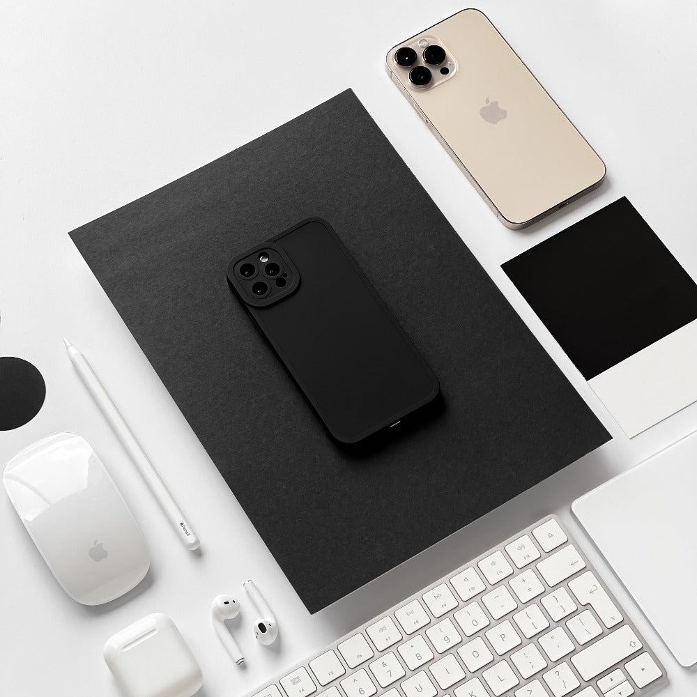 A flat lay image featuring various Apple products, including a MacBook in a black case, an iPhone with a sleek Coconut Lane NAKD Phone Case in Midnight Black offering full camera protection, an Apple mouse, a keyboard, AirPods, and an Apple Pencil. The items are neatly arranged on a white surface with black and white color swatches.