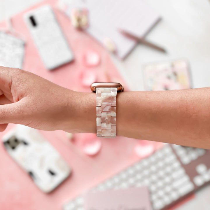 A person wears the Luxe Rose Apple Watch Strap by Coconut Lane, featuring a white, pearlescent, adjustable band on their wrist. The background displays a keyboard, notebooks, pens, and other office supplies, all in pastel colors and neatly arranged. Small pink hearts are scattered around, adding a decorative touch.