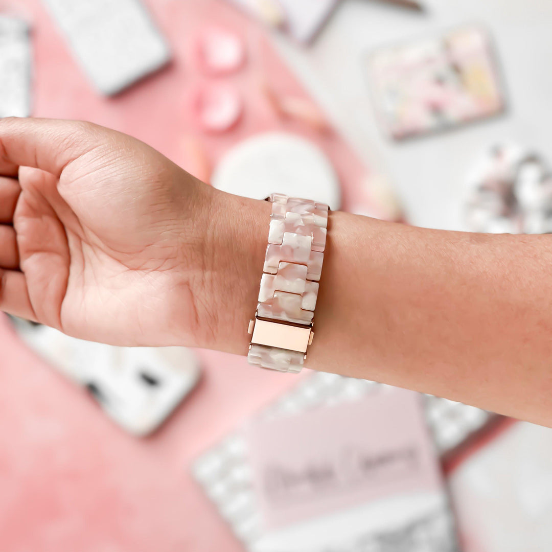 A person wears the Luxe Rose Apple Watch Strap by Coconut Lane, featuring a pink and white marbled, adjustable watch band. The background includes blurred objects in soft colors, such as notepads and desk items, creating an overall calm and aesthetic vibe.