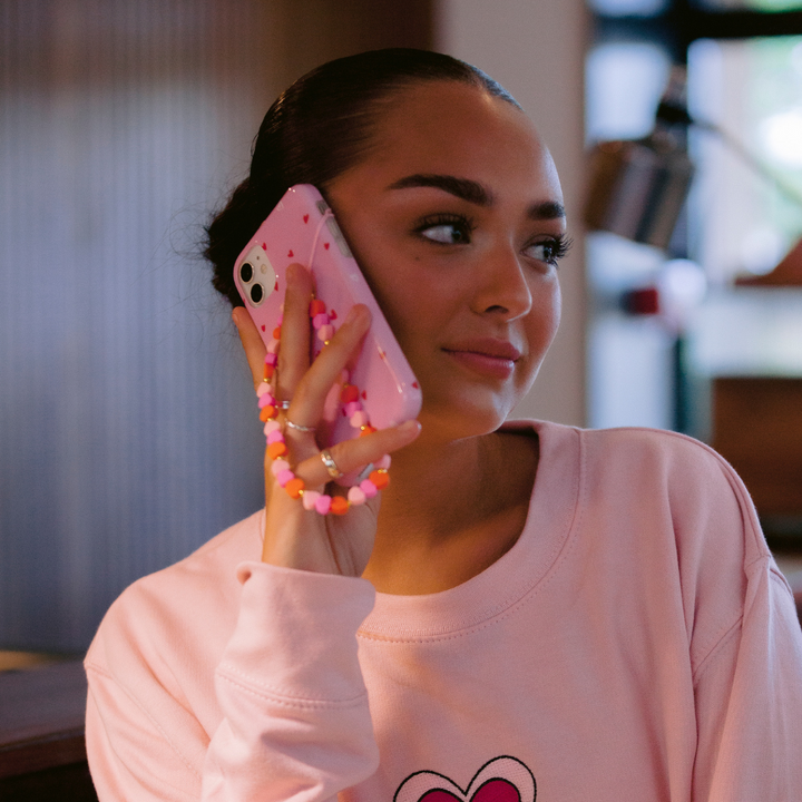 A person with a slicked-back hairstyle is holding a pink phone adorned with Coconut Lane's Beaded Phone Strap - Hearts to their ear while wearing a matching pink long-sleeve shirt. The vibrant phone charm adds a playful touch in the well-lit indoor setting, accentuating the blurred background.
