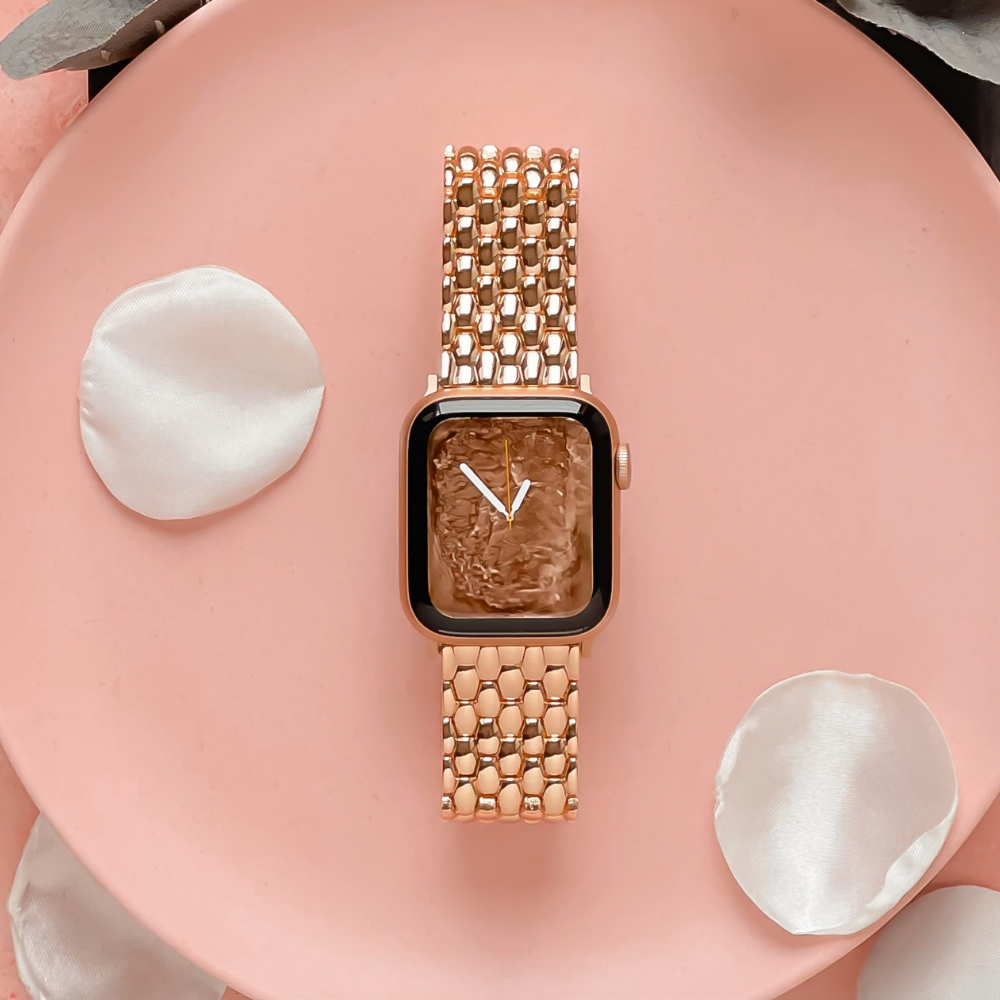 The Coconut Lane Mermaid Watch Strap in Rose Gold is beautifully showcased on a pale pink plate, adorned with three scattered white rose petals. The smartwatch screen features a minimalist white time display set against a textured background. Elegantly arranged, this setup also includes a complimentary link remover tool for effortless adjustments.