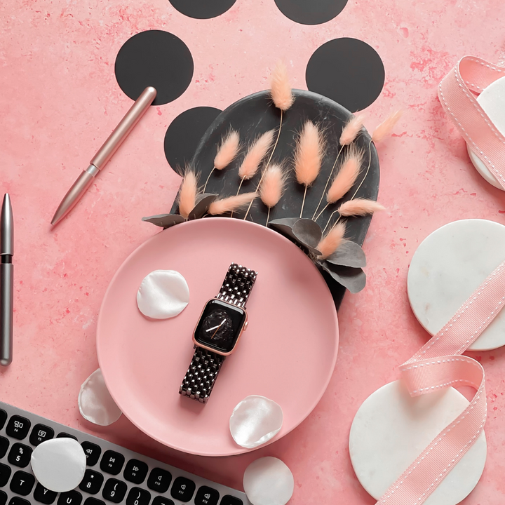 A flat lay on a pink textured surface featuring the Coconut Lane Mermaid Apple Watch Strap in black with a checkered band on a pink plate, surrounded by white petals, a corner of a black keyboard, a silver pen, and decorative dried flowers in a gray vase. Ribbons and marble coasters are also present.