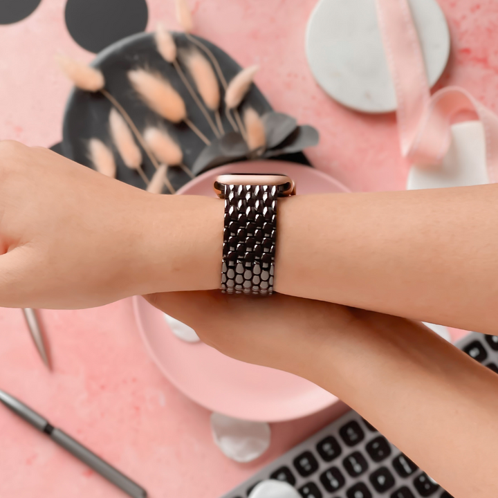 A person wearing a metallic wristwatch with an adjustable chain-link band is holding their wrist and adjusting the watch. The background features a keyboard, desk, pen, pink plate, pampas grass on a black plate, and a Coconut Lane Mermaid Apple Watch Strap - Black atop a pink surface.