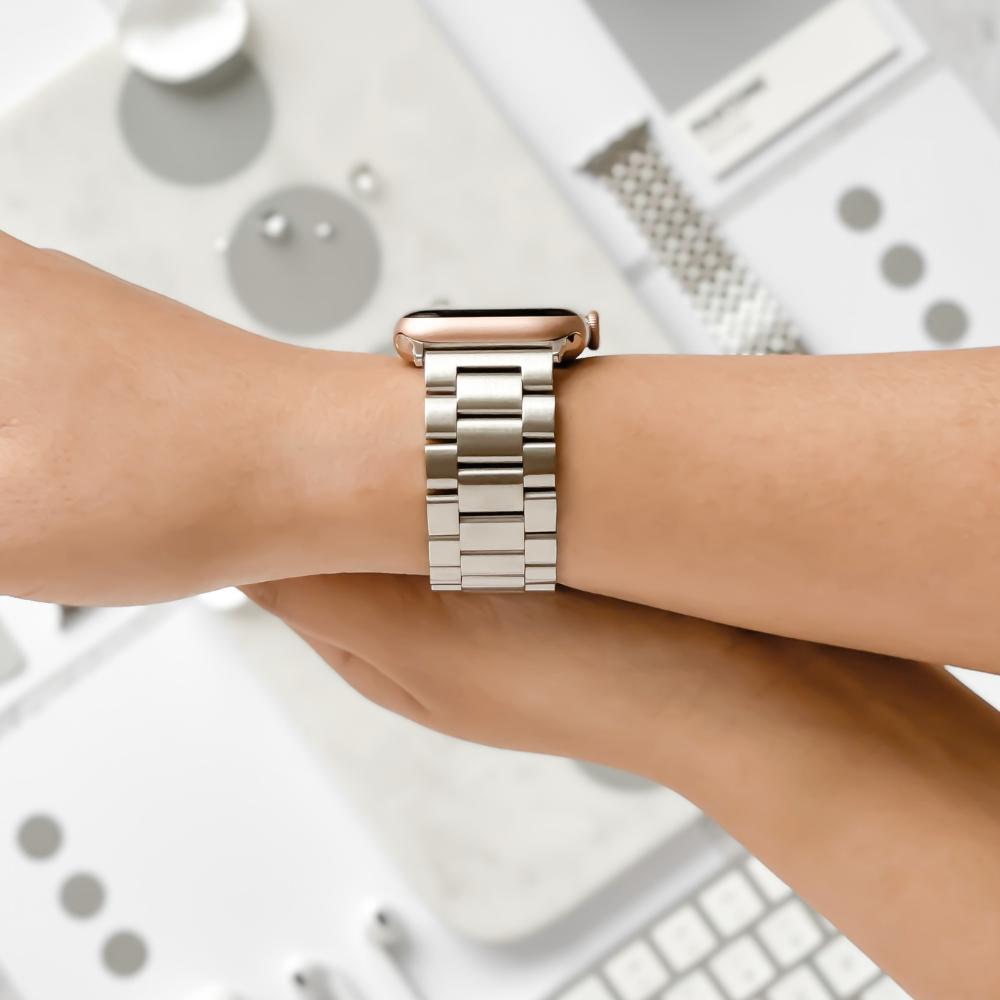 A person wearing the Coconut Lane Stainless Steel Apple Watch Strap in Silver on their wrist is seen. The background contains a blurred assortment of grey and white objects, including some watch accessories and tech gadgets. The focus is on the Apple Watch strap and the wrist.