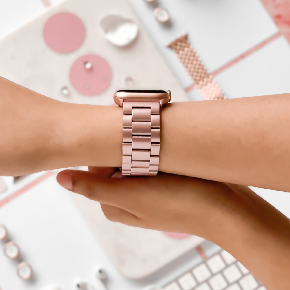 A close-up of a person's wrist wearing the Coconut Lane Stainless Steel Apple Watch Strap in Blush. The person is adjusting the strap with their other hand. The background features blurred desk items in pastel colors, including a keyboard, mouse, and stationery.
