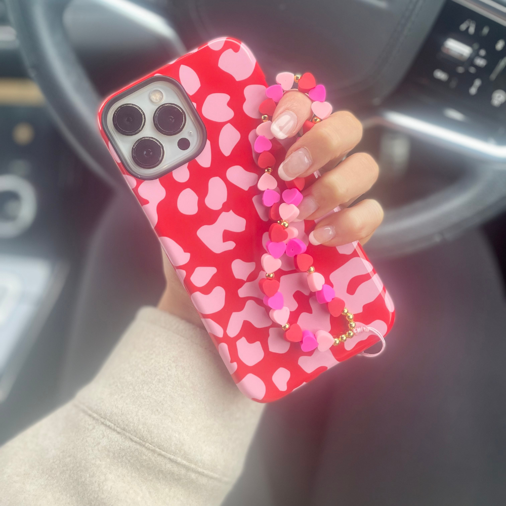 A person holding a smartphone with a red and pink patterned case is adorned with the "Beaded Phone Strap - Hearts" by Coconut Lane, featuring a heart-shaped bead charm. The phone is held in front of a car steering wheel, with the dashboard partially visible in the background.