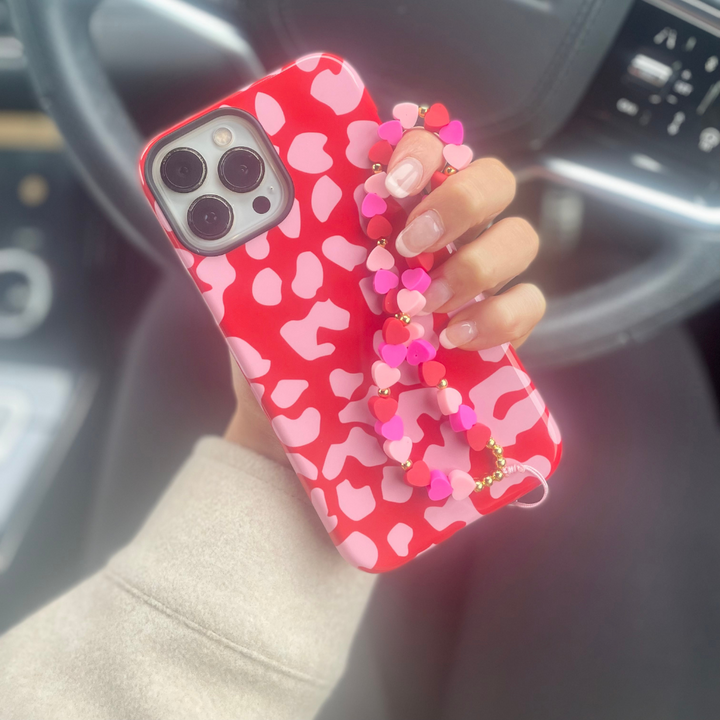 A person holding a smartphone with a red and pink patterned case is adorned with the "Beaded Phone Strap - Hearts" by Coconut Lane, featuring a heart-shaped bead charm. The phone is held in front of a car steering wheel, with the dashboard partially visible in the background.
