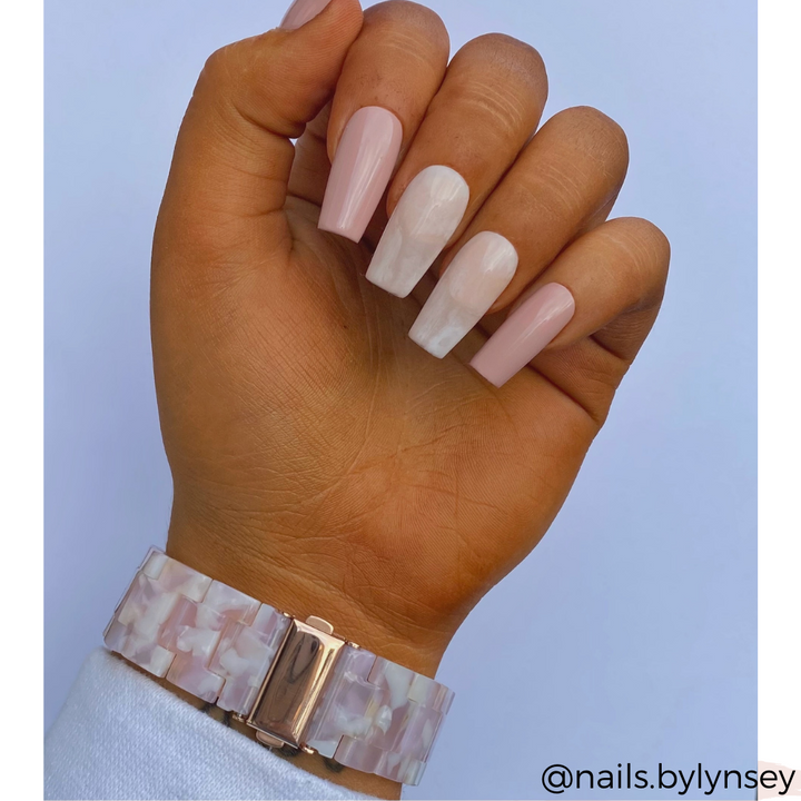 A hand with long, manicured nails; three nails are adorned with a light pink polish, while two showcase a marble-like white and pink design. The wrist sports the Luxe Rose Apple Watch Strap from Coconut Lane, featuring a matching pink and white marbled pattern. The text "@nails.bylynsey" appears in the bottom right corner.