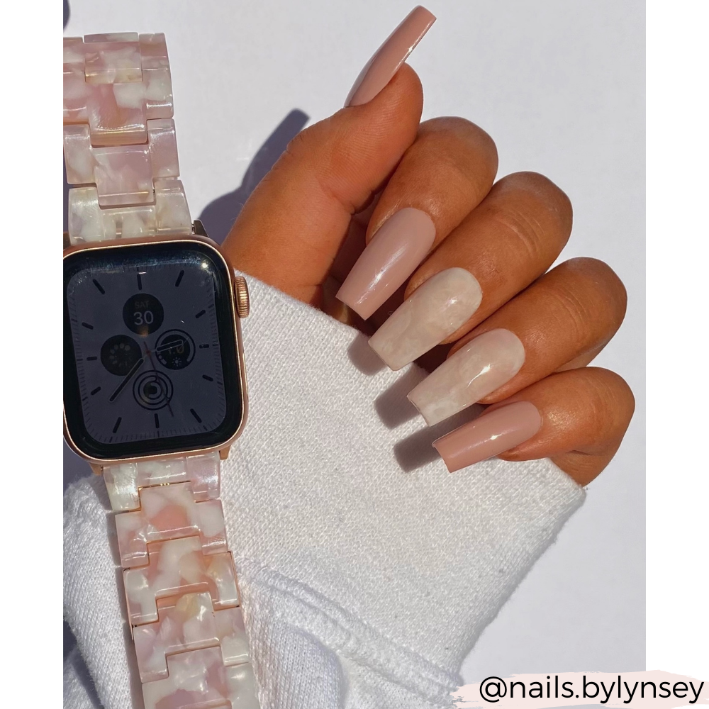A hand with long, coffin-shaped nails painted in a glossy neutral color, showcasing two accent nails featuring a marble design. The wrist is adorned with Coconut Lane's Luxe Rose Apple Watch Strap, which features an adjustable, light pink and white marbled pattern. The Instagram handle @nails.bylynsey is positioned at the bottom right.